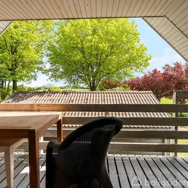 Træbord med en stol står på en overdækket terrasse. Udsigt til grønne og røde træer i en solrig have.