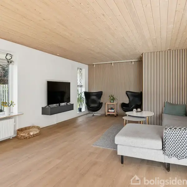 Stue med grå sofa, rundt sofabord og to sorte lænestole, placeret ved en trævæg. Et fladskærms-tv hænger på væggen. Store vinduer giver udsigt til en have.