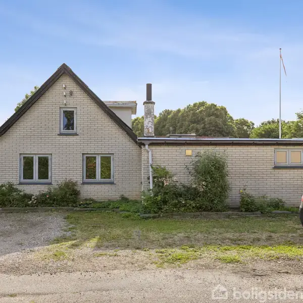 Et hvidt murstenshus med skrå tag hælder opad, omgivet af buske og træer. En bil holder parkeret foran huset på en grusvej under en klar blå himmel.