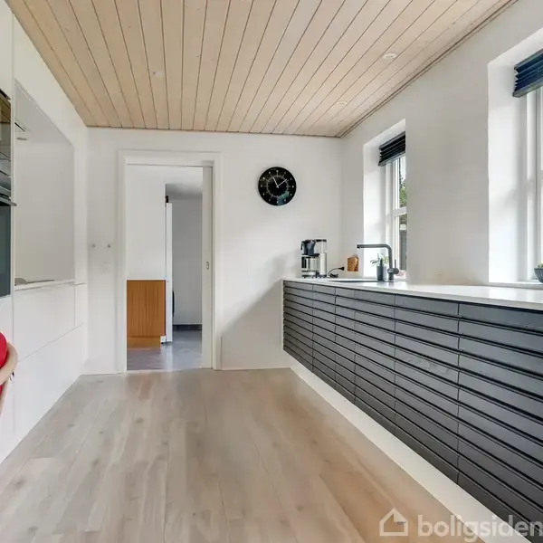 Køkkenbord med sorte skuffer under et hvidt bordplade med kaffemaskine. Vinduet viser et grønt landskab udenfor. Rød stol i venstre side og vægur på væggen.
