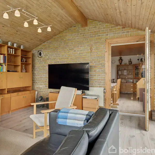 Stue med sort sofa og træstole omgivet af reoler med bøger og pynt. TV på murstensvæg. Skydedør åbner til spisestue med træmøbler. Loftlampe af træ.