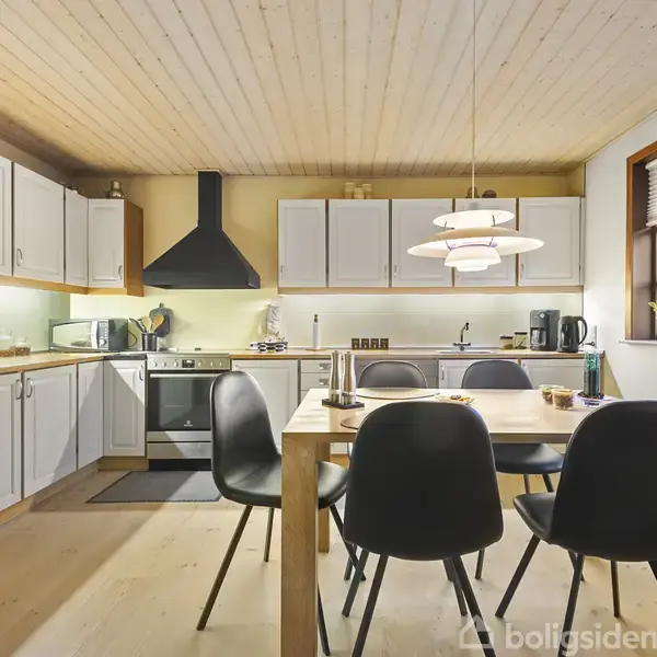 Et moderne køkken med hvide skabe, sort emhætte, og træbord omringet af sorte stole. Bordet er pyntet med lys, og vinduet giver naturligt lys. Lamper hænger over bordet.