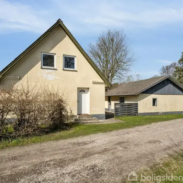 Et gult hus med to vinduer står ved en grusvej. I baggrunden er en mindre bygning og træer, der skaber en landlig atmosfære.