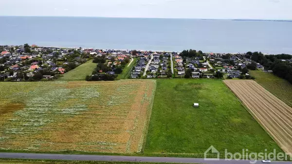 Grønne marker strækker sig mod række af huse ved kysten. Havet ligger i baggrunden under en overskyet himmel. En smal vej adskiller markerne fra husene.