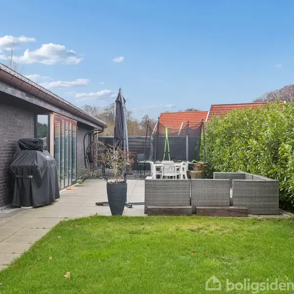 En terrasse med grill og pladestole står ved siden af et sort murstenshus. En grøn græsplæne omgiver området, med en høj hæk som baggrund under en klar blå himmel.