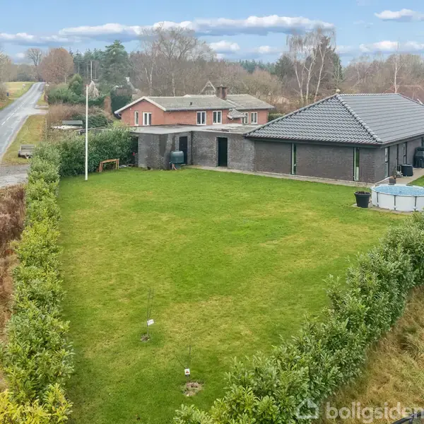 Hus med mørkt tag står ved en vej omgivet af en stor, velplejet græsplæne med buske omkring. Et andet hus ses længere væk, og vejen løber parallelt med ejendommen.