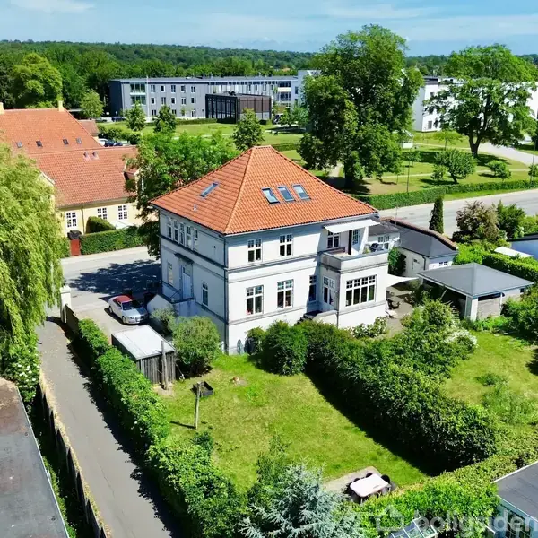 En hvid to-etagers villa med rødt tag står i en grøn have omgivet af buske og træer. Andre huse og en bygning ses i baggrunden under en klar blå himmel.
