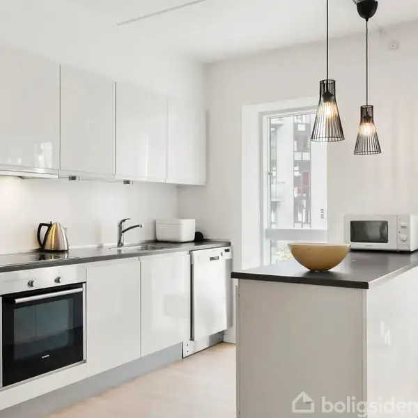 Et moderne køkken med hvide skabe og sort bordplade. En elkedel og skål står på bordet. Hængelamper over bordpladen, vindue i baggrunden, plante i hjørnet.