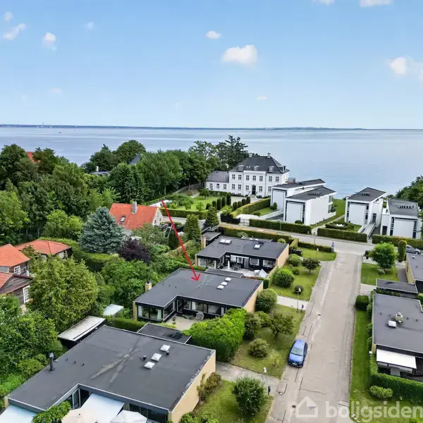 Et parcelhus (markeret med rød pil) ligger i et grønt boligområde ved vandet med moderne og traditionelle bygninger omkring. En vej adskiller husene, og havet ses i baggrunden.