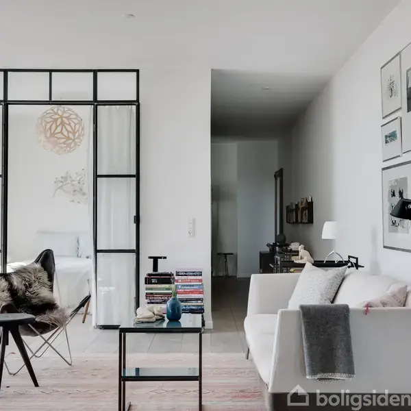Stue med hvid sofa, sofabord og lænestol med fåreskind, adskilt fra et andet rum med skydedøre i glas. Væggene er dekoreret med forskellige rammer og billeder.