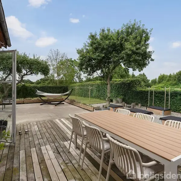 En træterrasse med et langt bord og otte stole står foran et hus. En hængekøje er placeret i haven, omgivet af grønne buske og træer under en blå himmel.