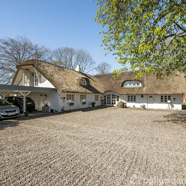 Et hvidt bindingsværkshus med stråtag står stille i en stor gårdsplads omgivet af træer. Til venstre er en carport med en parkeret bil. Solrigt vejr.