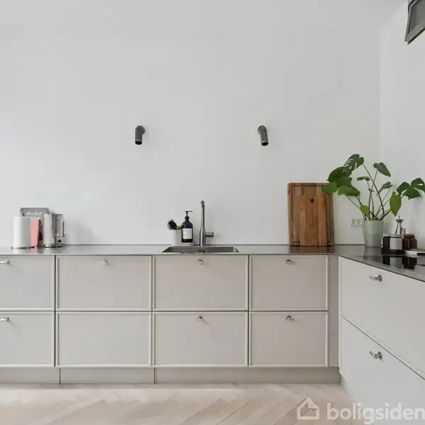 Køkken med hvid skabsrække, stålvask, kaffemaskine og skærebræt. Grøn plante ved siden af vasken. Højre side har indbygget ovn. Hvid væg og lyse trægulve omgiver.