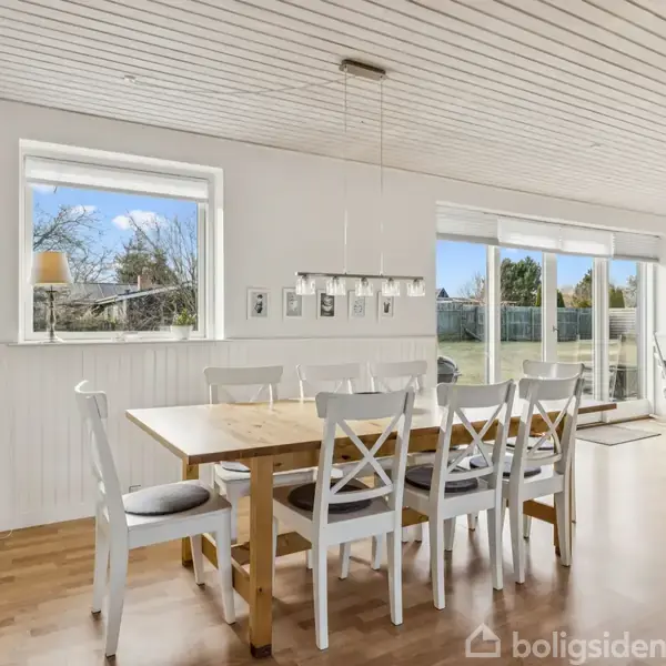 Et spisebord med hvide stole står i en lys stue. Store vinduer giver udsigt til en have, og en sofa ses i baggrunden. Pragmatisk indrettet og lys atmosfære.