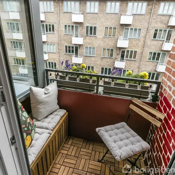 En lille balkon med en bænk og en stol med hynder, omgivet af en murstensvæg. Den har udsigt til en murstensbygning med flere vinduer og altaner.