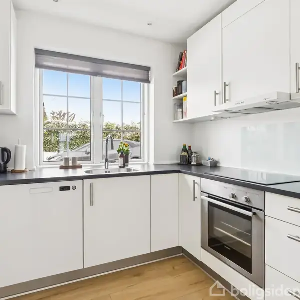 Et lyst køkken med hvide skabe og sort bordplade. En ovn og kogeplade er indbygget, kaffemaskine og elkedel står på bordet. Et vindue med udsigt til buske.