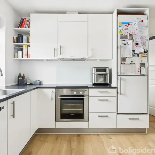 Et hvidt køkken med moderne skabe, indbygget ovn og mikrobølgeovn, køleskab fyldt med tegninger. Der er en vask med vandhane ved vinduet og træplankegulv i rummet.