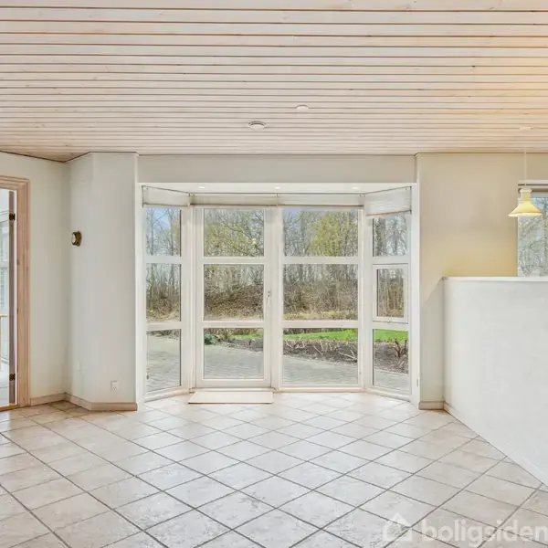 Stort, lyst rum med flisegulv og store vinduer, der skaber udsigt til en skov. Spisestue til venstre med træmøbler gennem en åben dør. To hængelamper til højre.