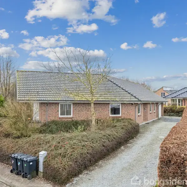 En murstensvilla med grå tagsten står stille omgivet af en grusindkørsel og hække, under en blå himmel med spredte skyer; i nærheden er der skraldespande ved vejkanten.