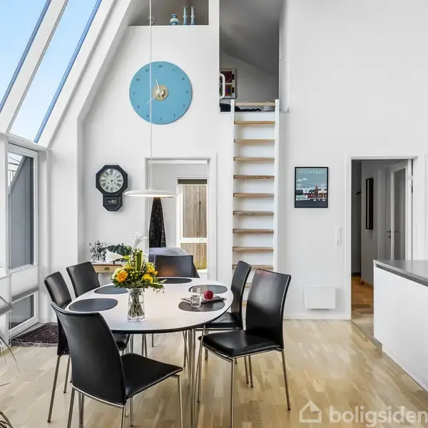 Et moderne spisekøkken med et rundt bord og sorte stole. Sollys strømmer ind gennem store skråvinduer. En blå vægur og en stige til en hems ses i baggrunden.