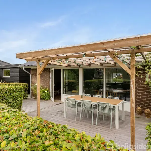 Terrasse med træpergola har et langbord og stole, omgivet af grønne buske og en murstensbygning i baggrunden. Solen skinner, hvilket indikerer en klar dag.
