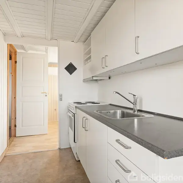 Et køkken med hvide skabe og skuffer står stille. Bordpladen er grå med en indbygget vask. Væggene og dørene er lyse, og loftet har hvidmalede bjælker.