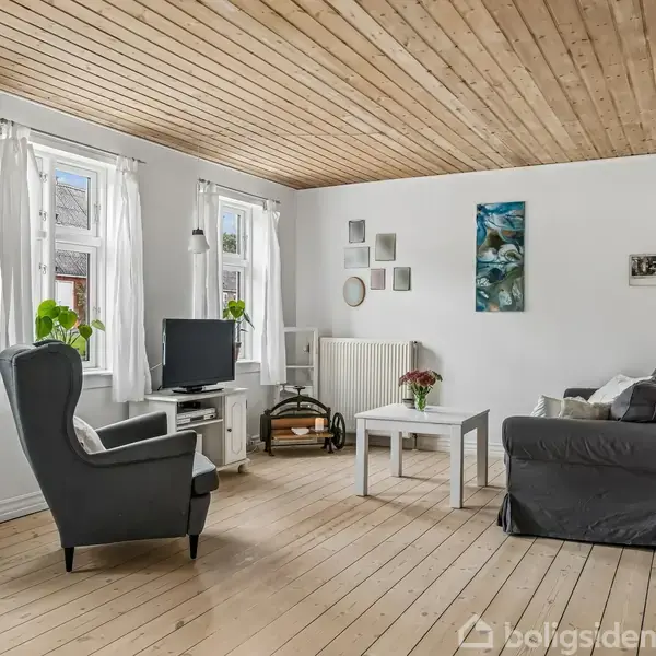 En stue med en grå lænestol og sofa omkring et hvidt sofabord. Fjernsynet står ved et vindue. Trægulv og loft skaber en lys og enkel atmosfære.