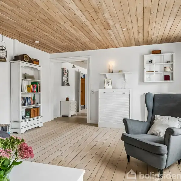 Grå lænestol står i en lys stue med trægulv og loft, omgivet af en reol med bøger og pyntegenstande. Et hvidt sofabord med blomster står i forgrunden.