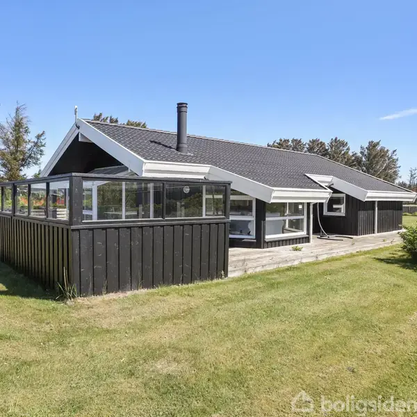 Et sortmalet sommerhus med glasindhegnet veranda står på en velplejet græsplæne. Omkranset af træer, under en blå himmel, med en skorsten øverst på taget.