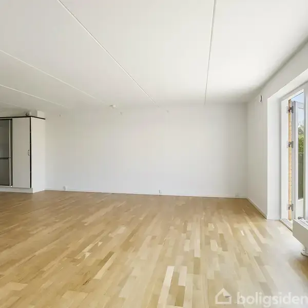 Stue med trægulv åbner op mod køkken til venstre. Store vinduer til højre giver udsigt til en terrasse og have. Rummeligt med hvide vægge og naturligt lysindfald.