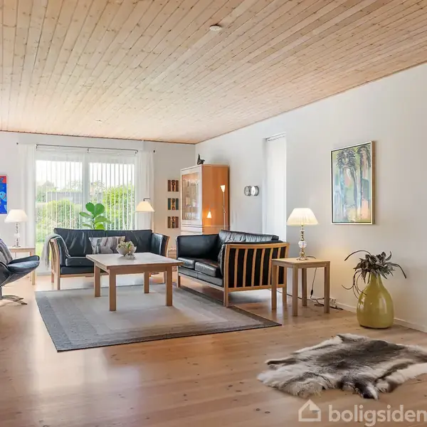 Stue med sorte lædersofaer og træmøbler, placeret omkring et sofabord. Lamper og malerier pynter rummet. Træloft og lyse gulve skaber en lys atmosfære. Grønne planter ved vinduet.