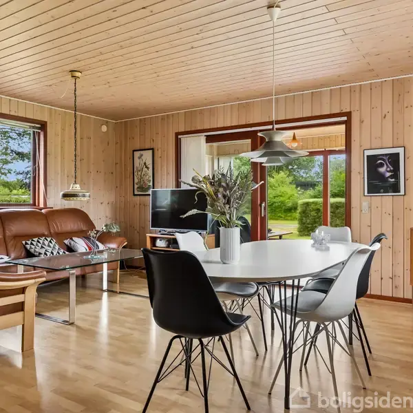 Stue med træbeklædte vægge, en lædersofa ved siden af et glaskaffebord, hvorpå en rund spisebord med sorte stole står. Store vinduer giver udsigt til en grøn have.