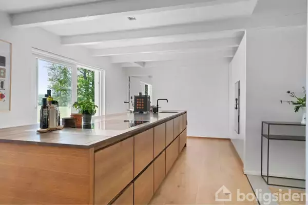 Køkkenø med træfinish og metaloverflade står i moderne, lys køkken, omgivet af vinduer med udsigt til grønne træer. Rummets hvide vægge og loftsbjælker skaber en stilren atmosfære.