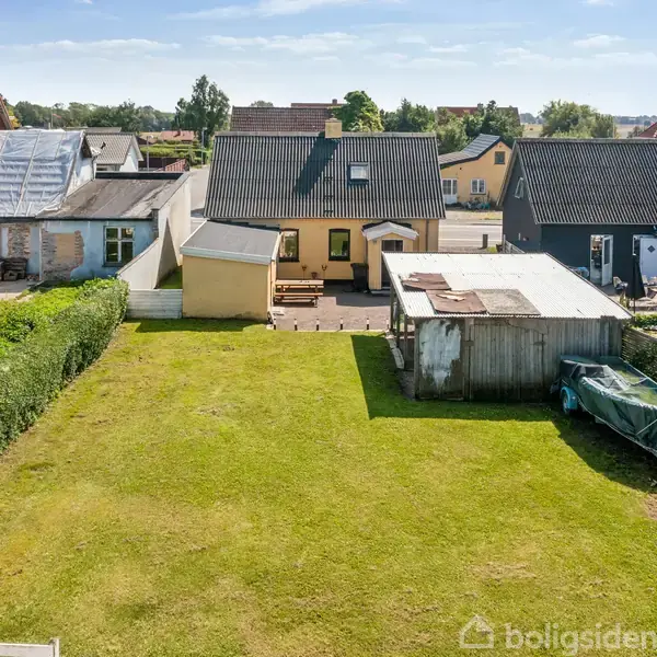 En græsplæne strækker sig mellem to rækker huse. Foran plænen står en trækonstruktion ved siden af et gammelt bådskrog. Hækken omkranser plænen. Baggrunden har flere huse.