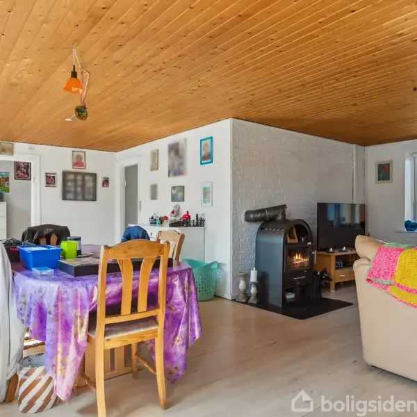 Et træbord dækket med en lilla dug står i et lyst rum med træloft, ved siden af en brun sofa og en brændeovn. Væggene er dekoreret med diverse billeder.