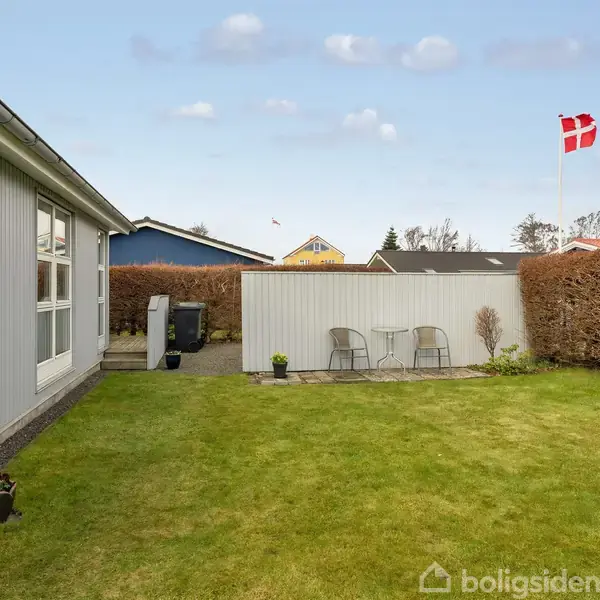Græsplæne omgivet af hække med en flagstang der bærer det danske flag. Til højre står to stole ved en hæk. Bygning til venstre med store vinduer.