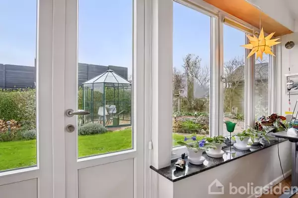 En glasdør fører til en have med en grøn græsplane og et drivhus. Indenfor står en række potteplanter på en vindueskarm. En stjerneformet lampe hænger i hjørnet.