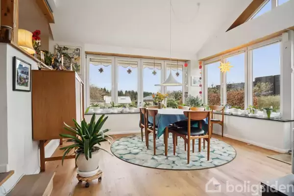 Spisebord med blå dug og fem stole står i en lys stue med store vinduer og udsigt til en have. Planter og dekorationer er placeret på vindueskarmen.