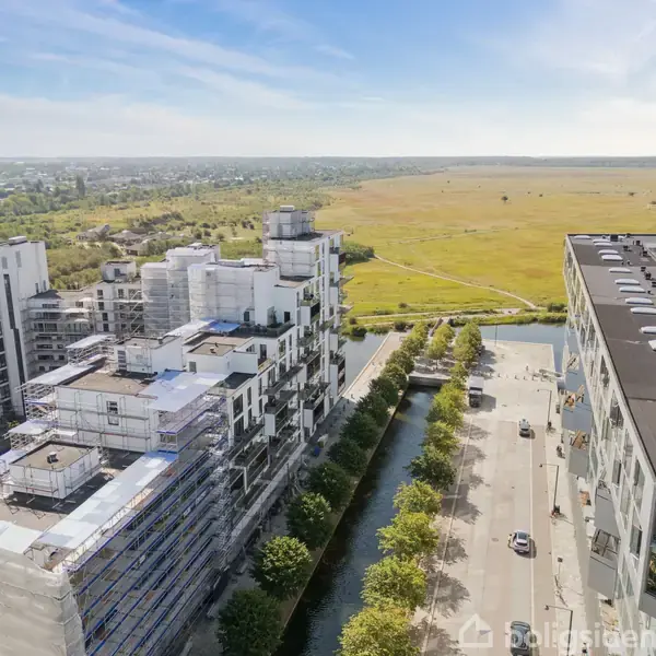 Flere moderne lejlighedskomplekser ligger på hver side af en gade med træer, der fører til åbne, grønne områder i horisonten under en klar himmel.