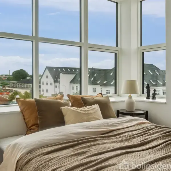 En seng med puder står foran store vinduer, der viser et udsigt til en bydel med hvide bygninger og blå himmel. På et bord ved siden af står en lampe og små figurer.