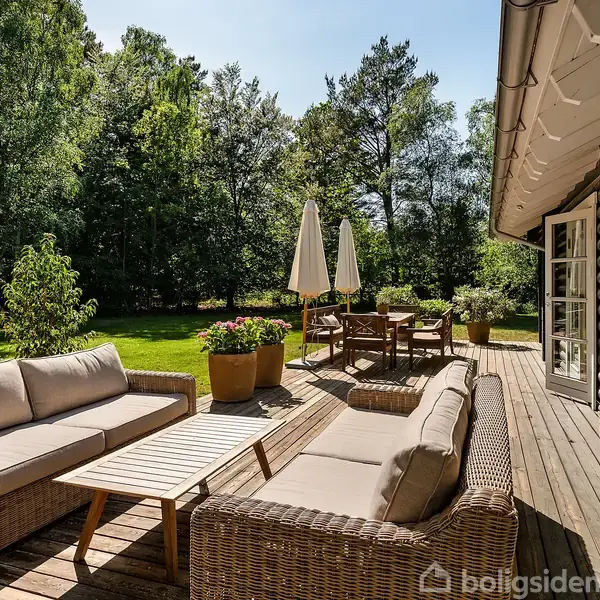 Terrasse med flettede møbler og sofabord, omgivet af grønne planter og træer. To parasoller står ved et spisebord. Skovklædt have skimtes i baggrunden.