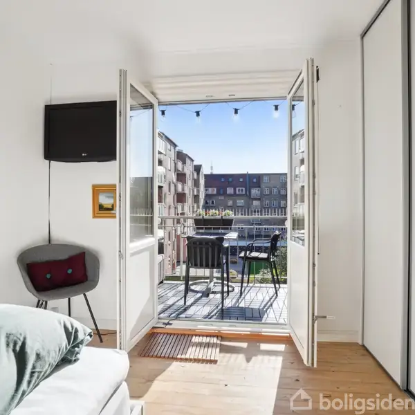 Soveværelse med en åben dobbeltdør fører ud til en altan med stole. Et ophængt fjernsyn og en stol ses til venstre, mens en stor skydedørsgarderobe er til højre.