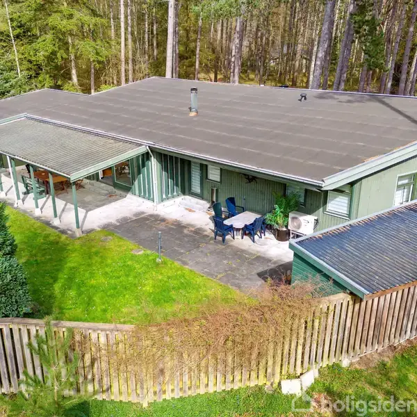 Hus med fladt tag står stille i skovområde, omkranset af træer og hegn. Foran huset er der en terrasse med havemøbler og en græsplæne.