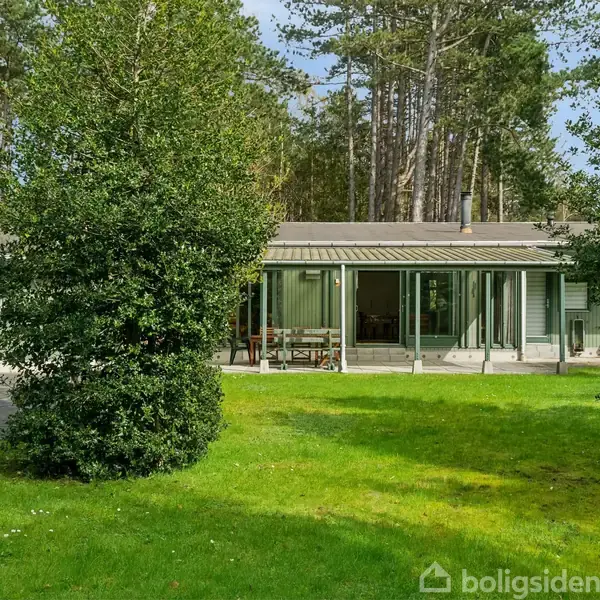 En enkeltstående, grøn bygning står stille i en frodig have med græsplæne, omgivet af tætte buske og høje træer i baggrunden, under en klar, blå himmel.