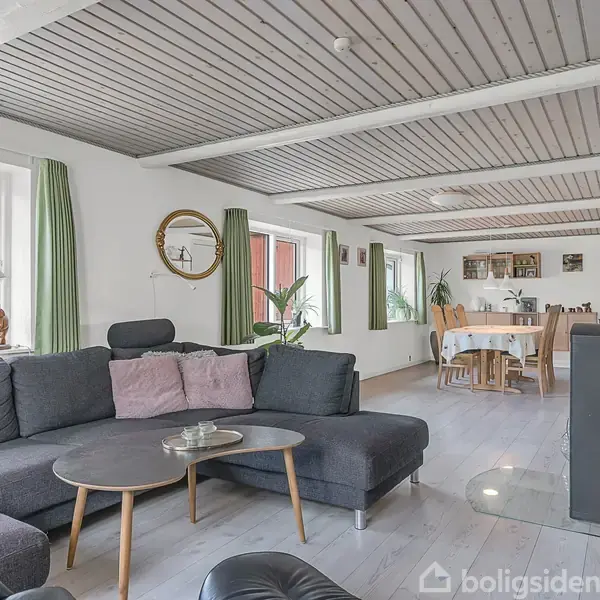 Stor, hyggelig stue med grå sofa og rund sofabord. Stuen har lyse trægulve og grønne gardiner. Der er en spiseplads med træbord i baggrunden, omgivet af vinduer og dekorative planter.