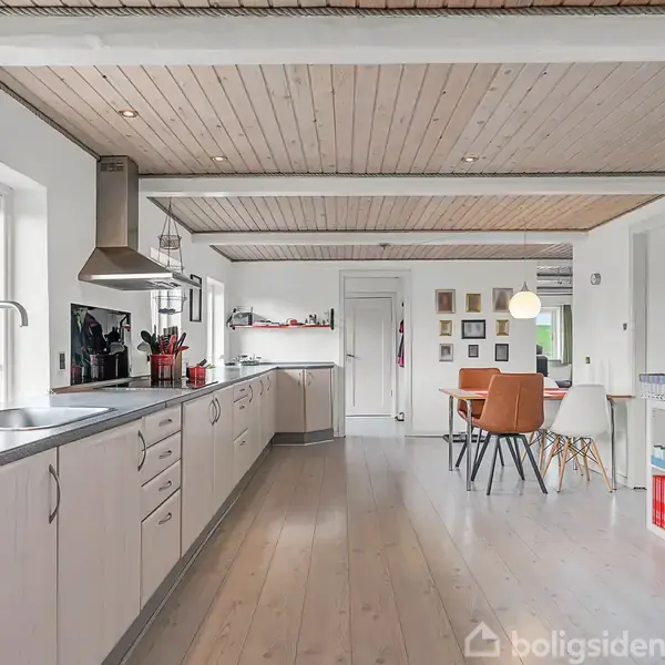 Et lyst køkken med træloft og lyse træplanker på gulvet. Lange, hvide køkkenelementer med indbygningsovn. Spisebord i baggrunden, store vinduer mod venstre med udsigt til grønne omgivelser.