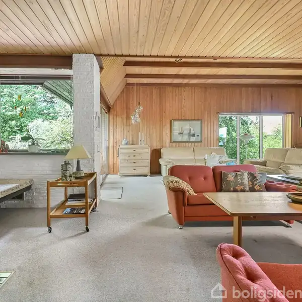 Stue med trælofter og vægge af skiftevis mursten og træ. Sofaer og bord i midten, store vinduer med udsigt til grønne omgivelser. Hylder med bøger langs væggen.