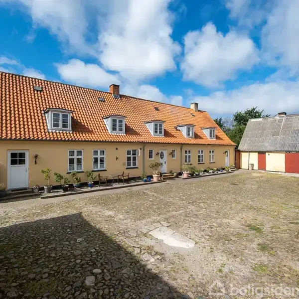 Et gult bondehus med rødt tegltag står stille i en gårdsplads omgivet af gårdbygninger på en solskinsdag med blå himmel og hvide skyer.