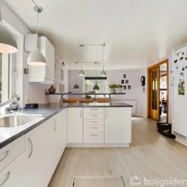 En hvidt køkken med moderne skabe og stålvaske. Hængende lamper lyser rummet op. Et stort vindue viser en have udenfor. En dør fører til en tilstødende stue.