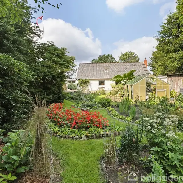 Hvidt hus med stråtag ligger i baggrunden omgivet af en frodig have med blomsterbede og et lille drivhus. Himmel med skyer og grønne træer udgør den omgivende udsigt.